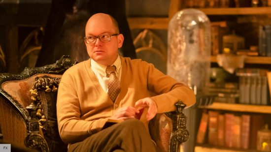What We Do In the Shadows cast and characters: Mark Proksch as Colin Robinson sitting in a chair in the vampire Rhode Island home